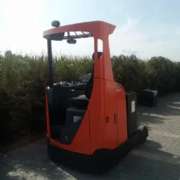 Pomarańczowy wózek widłowy elektryczny typu reach truck stoi na kostce brukowej, za nim widoczne krzaki i błękitne niebo.