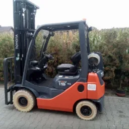 Pomarańczowo-szary wózek widłowy Toyota 15 SAS stojący na kostce brukowej z zaroślami w tle.