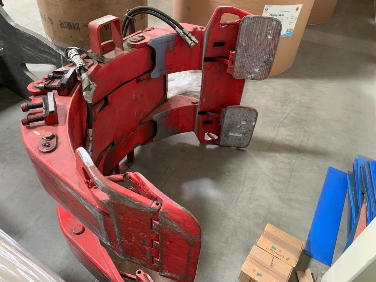 Czerwony, używany chwytak do wózka widłowego z hydraulicznymi przewodami, widoczne ślady użytkowania i otarcia farby, na tle betonowej podłogi magazynu.