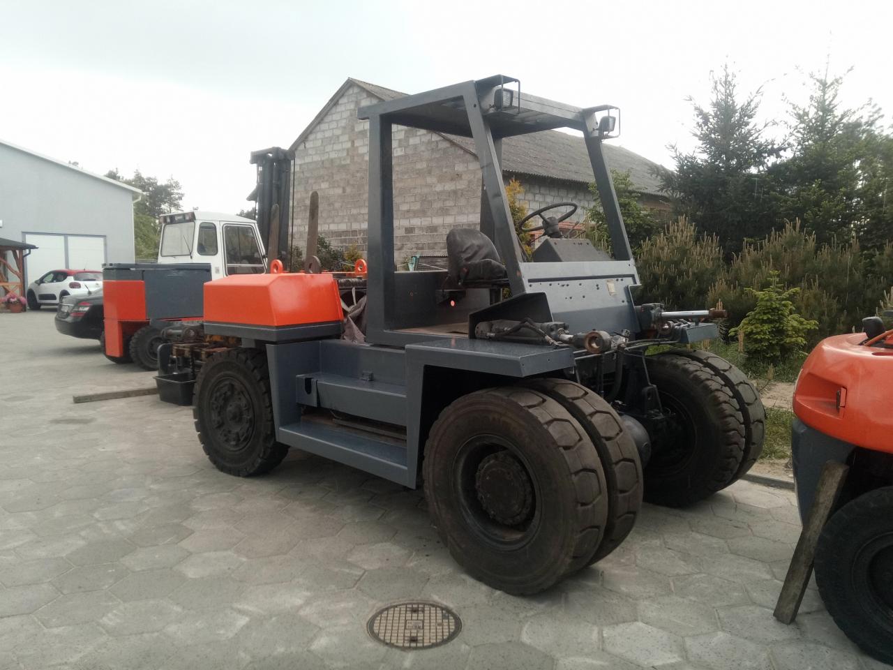 Szary wózek widłowy z kabiną i dużymi oponami, zaparkowany na brukowanym placu, w tle budynek gospodarczy