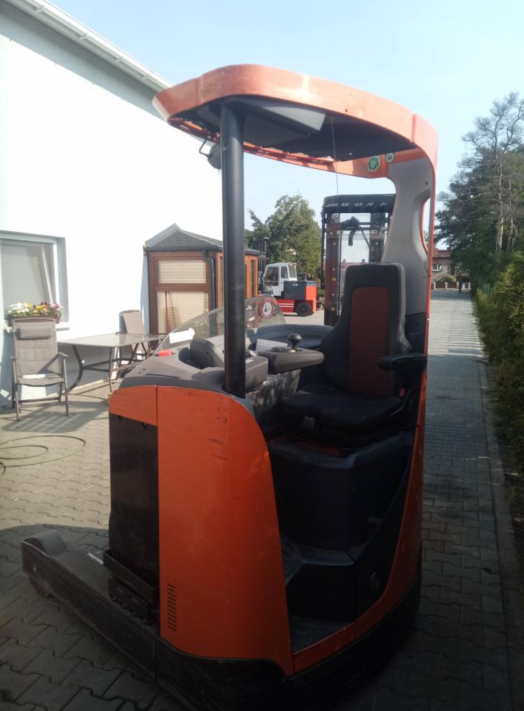 Pomarańczowo-szary wózek widłowy elektryczny stojący na zewnątrz, widok z boku z perspektywą na kabinę operatora, w tle budynek i drugi wózek widłowy.