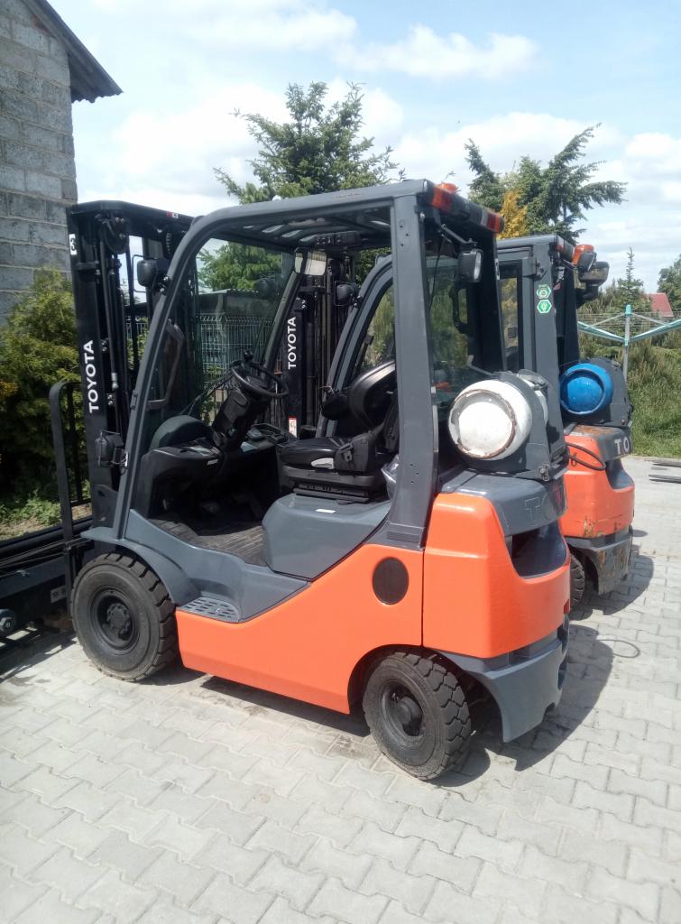 Dwa wózki widłowe Toyota w Godziesze Małe, jeden szary z pomarańczowym korpusem i butlą gazową, drugi pomarańczowy, stoją na kostce brukowej na zewnątrz.