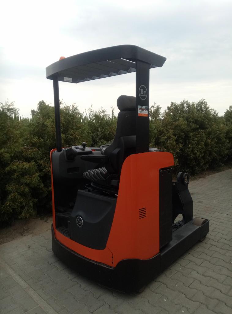 Pomarańczowo-czarny wózek widłowy elektryczny stojący na kostce brukowej, z zielonymi krzewami w tle i pochmurnym niebem.