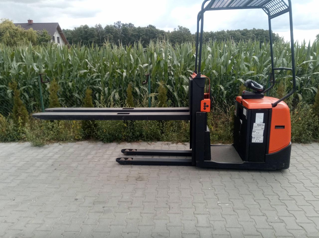 Elektryczny wózek paletowy z masztem, pomarańczowo-czarny, na kostce brukowej, z polem kukurydzy w tle.