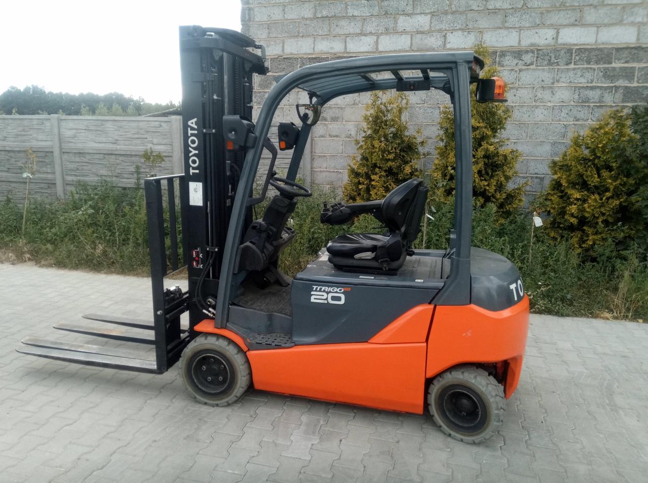 Pomarańczowo-szary wózek widłowy Toyota Trigo 20 na kostce brukowej, z widocznym logo marki i elementami konstrukcyjnymi, na tle muru i zieleni.