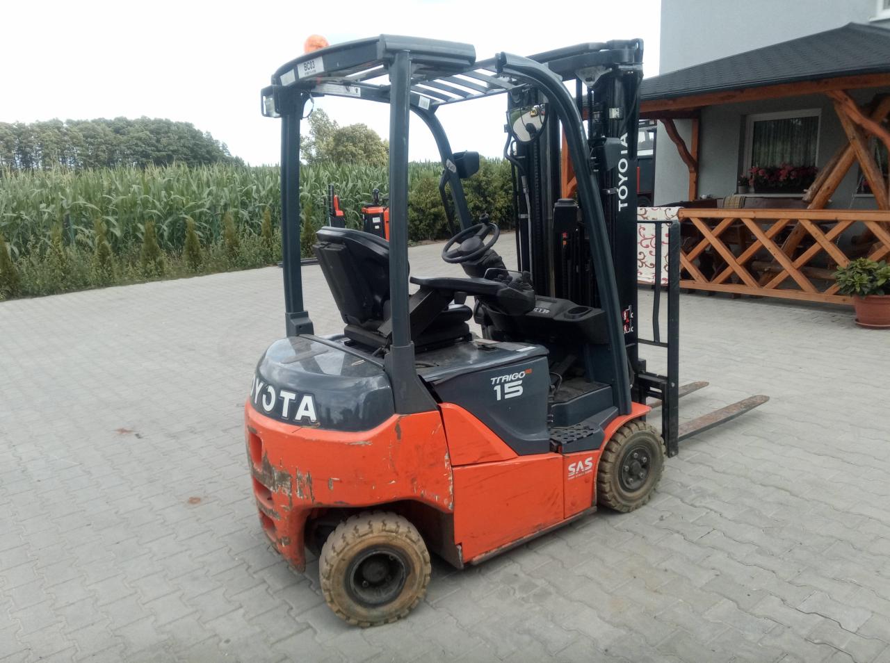 Używany wózek widłowy Toyota TTRAIGO 15, pomarańczowo-szary, stoi na kostce brukowej na tle pola kukurydzy i budynku z drewnianą konstrukcją.