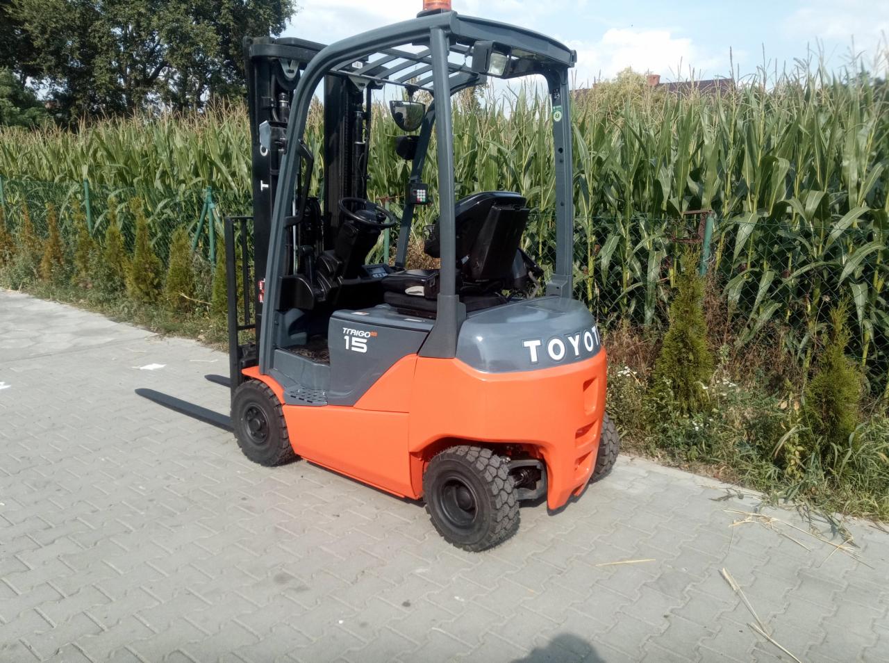 Pomarańczowo-szary wózek widłowy Toyota, model TFRIGO 15, stoi na kostce brukowej na tle pola kukurydzy.