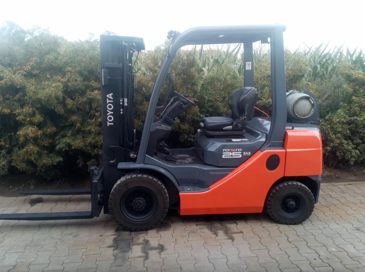 Pomarańczowo-szary wózek widłowy Toyota Tonero 25 SAS z butlą gazową na tle zielonych krzewów i pola kukurydzy.