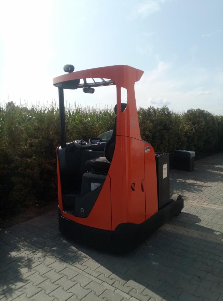 Pomarańczowy wózek widłowy elektryczny typu reach truck stoi na kostce brukowej, za nim widoczne krzaki i błękitne niebo.