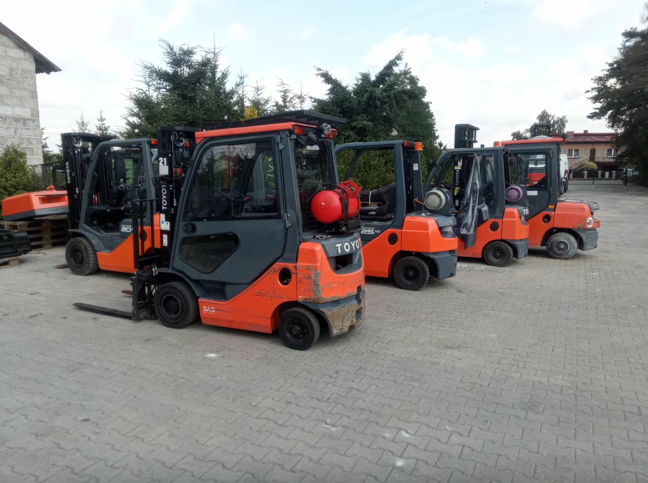 Rząd pomarańczowo-szarych wózków widłowych Toyota na placu, jeden z czerwoną butlą gazową, pozostałe z szarymi, widoczne ślady użytkowania.