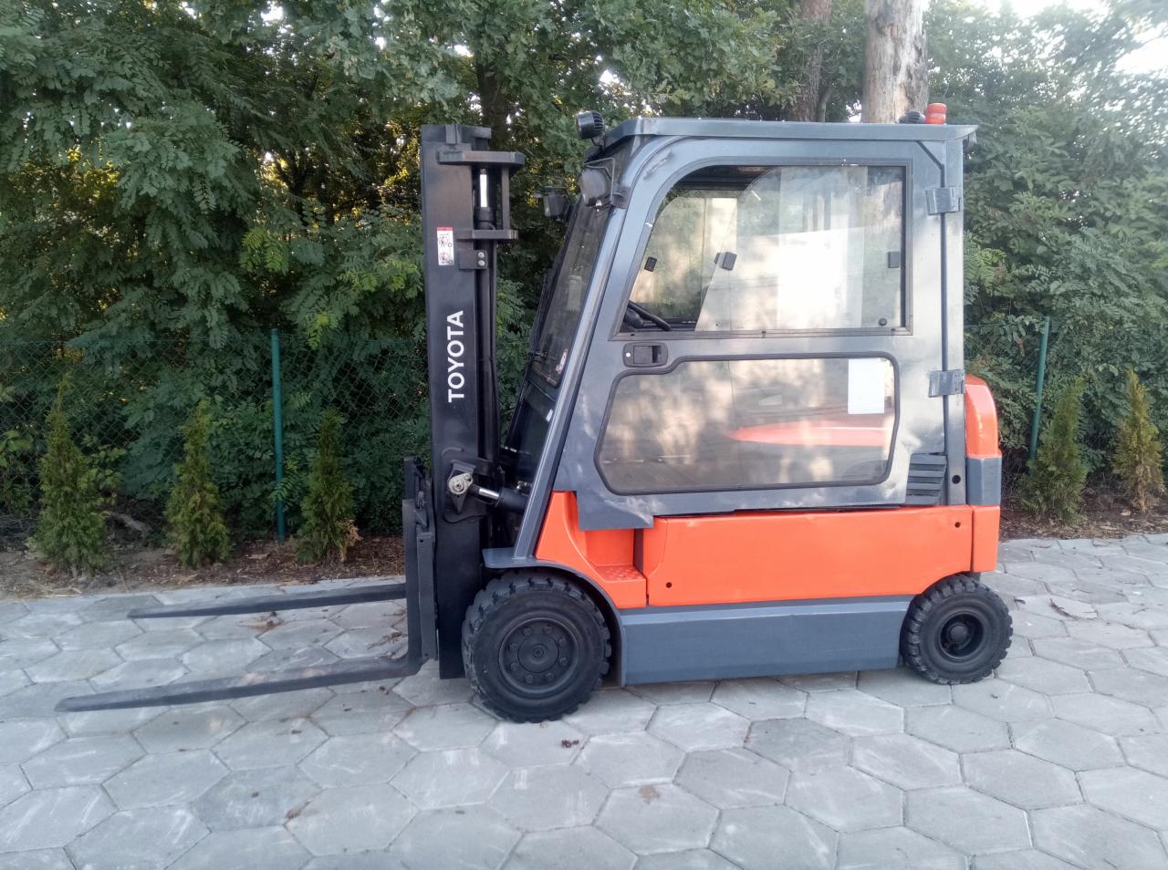 Nowy, pomarańczowo-szary wózek widłowy Toyota na kostce brukowej, z zielenią w tle, widok z boku.