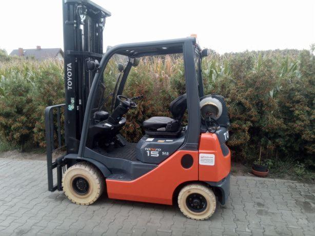 Pomarańczowo-szary wózek widłowy Toyota 15 SAS stojący na kostce brukowej z zaroślami w tle.