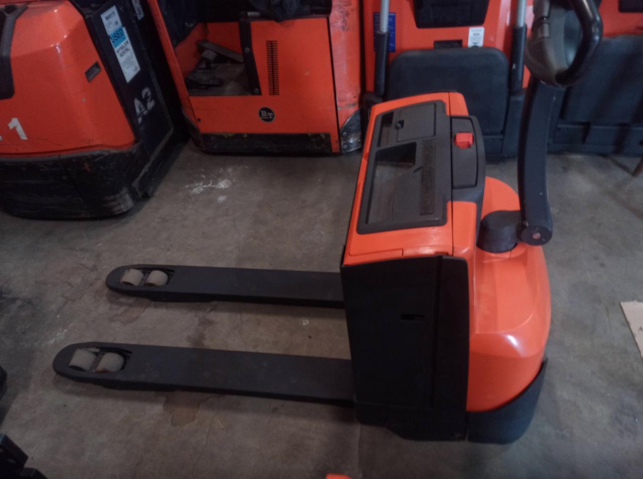 Pomarańczowo-czarny, elektryczny wózek paletowy z widłami, stojący na betonowej podłodze magazynu, z innymi wózkami w tle.