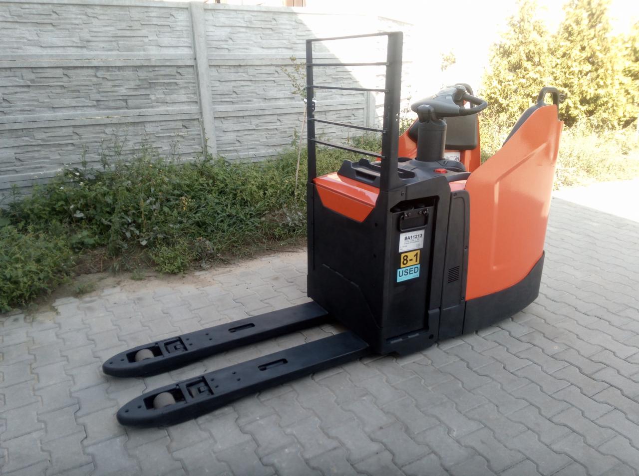 Pomarańczowo-czarny wózek paletowy elektryczny z naklejką 'USED' na tle betonowego ogrodzenia i kostki brukowej.