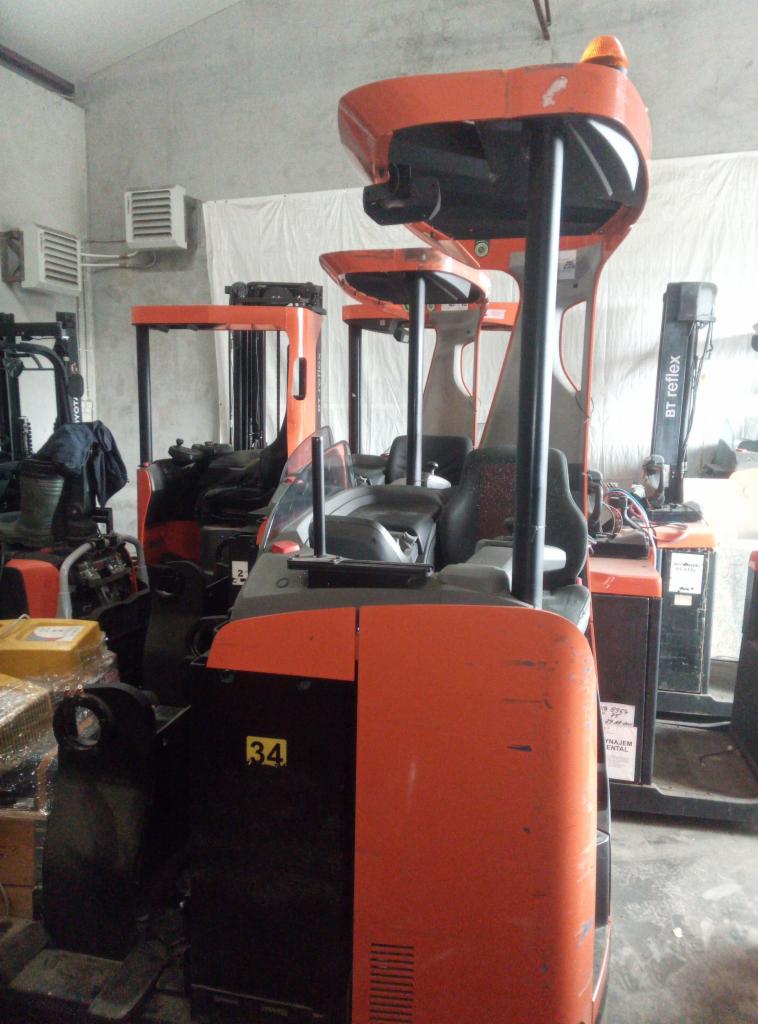 Pomarańczowy wózek widłowy elektryczny z numerem 34 na obudowie, stojący w magazynie z innymi wózkami.