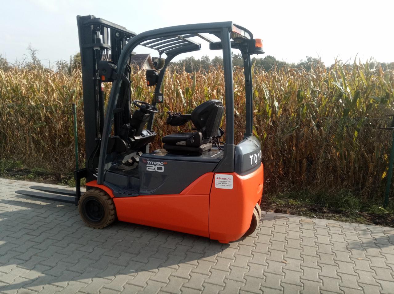 Nowy, pomarańczowo-szary wózek widłowy Toyota Trigo 20 na kostce brukowej, z polem kukurydzy w tle.