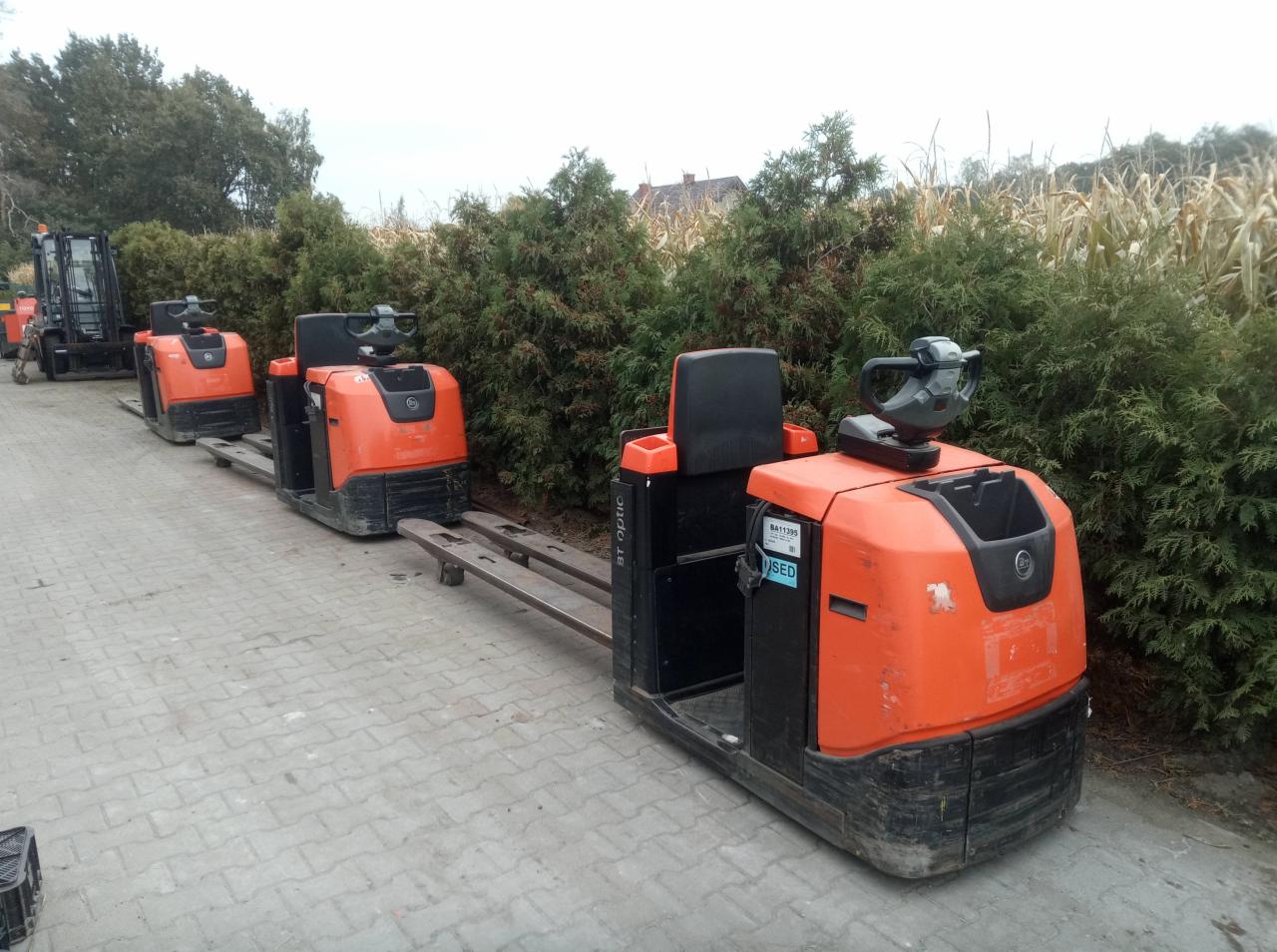 Rząd pomarańczowych wózków paletowych elektrycznych BT Optio na zewnątrz, z widoczną naklejką 'USED' na jednym z nich, na tle zielonych krzewów i kukurydzy.