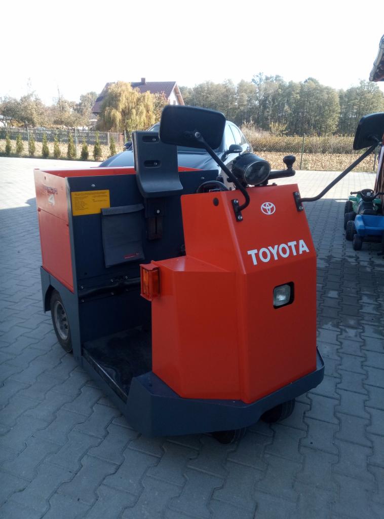 Pomarańczowo-granatowy wózek transportowy Toyota na kostce brukowej, widoczne lusterka, lampa i małe koła, w tle dom i drzewa.