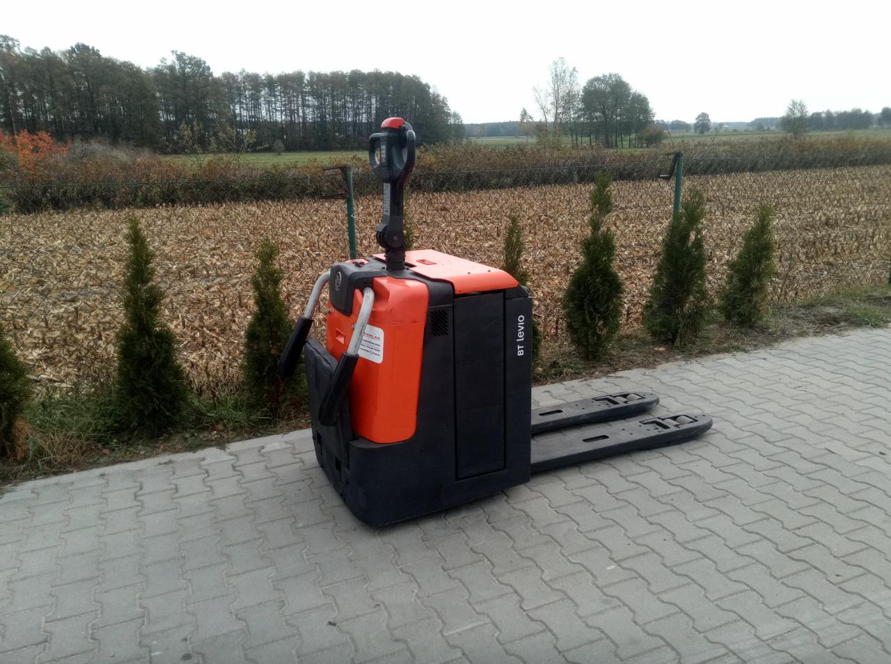 Pomarańczowo-czarny elektryczny wózek paletowy BT Levio stoi na kostce brukowej, na tle pola i drzew.