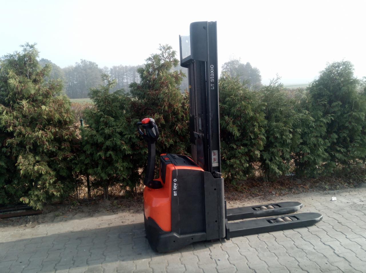 Pomarańczowo-czarny wózek widłowy elektryczny BT Levio O stoi na kostce brukowej, z żywopłotem z tui w tle.