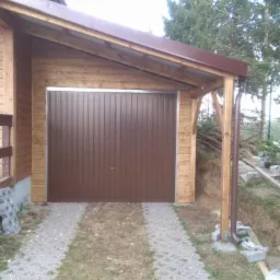 Zadaszone miejsce postojowe z brązową bramą garażową, drewnianą konstrukcją i częściowo ułożoną nawierzchnią z ażurowych płyt betonowych.