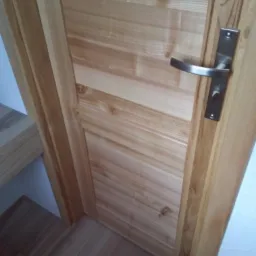 Jasne, drewniane drzwi wewnętrzne z panelem i metalową klamką, widoczne fragmenty drewnianej podłogi i schodów.