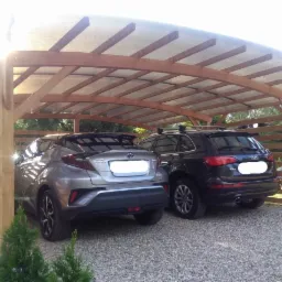 Drewniana konstrukcja z zadaszeniem z poliwęglanu, służąca jako wiata dla dwóch samochodów, stojąca na żwirowym podłożu, z widocznymi elementami ogrodzenia w tle.