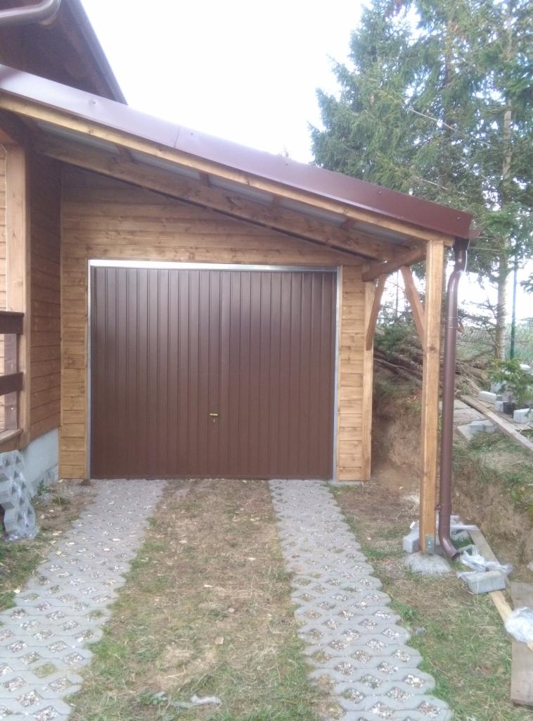Zadaszone miejsce postojowe z brązową bramą garażową, drewnianą konstrukcją i częściowo ułożoną nawierzchnią z ażurowych płyt betonowych.