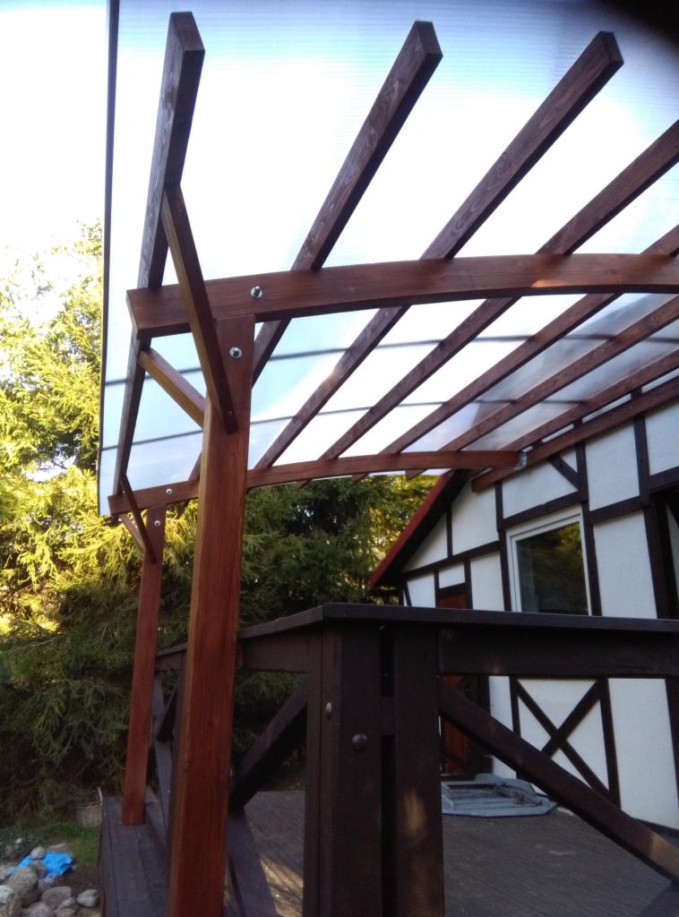 Zadaszenie tarasu z brązowych belek drewnianych i przezroczystego poliwęglanu, widok z dołu, z fragmentem domu w tle.