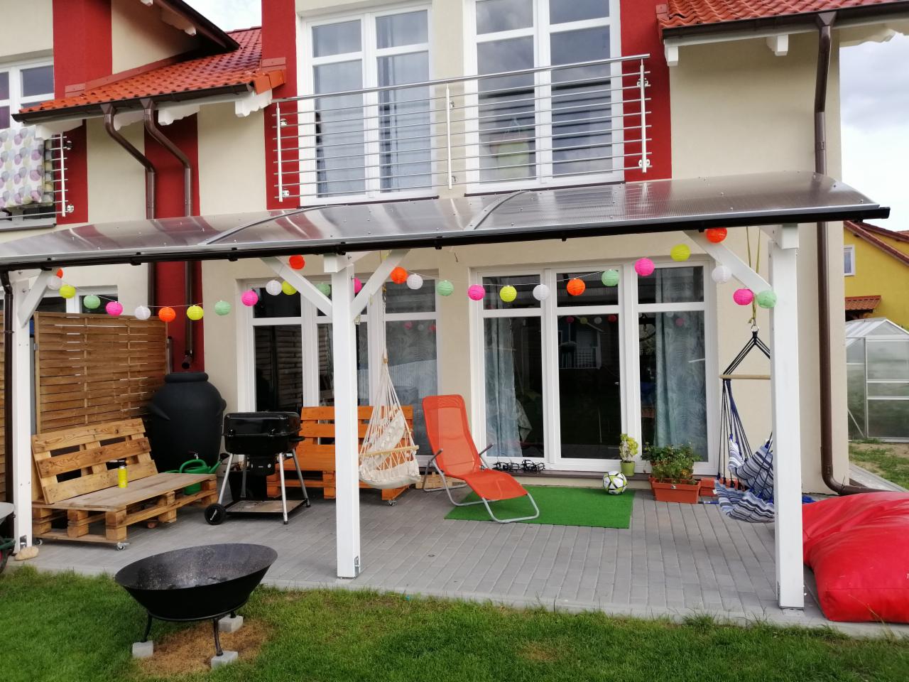Zadaszony taras z białymi słupami, ozdobiony kolorowymi lampionami, z meblami ogrodowymi, grillem i miejscem na ognisko, na tle dwupiętrowego domu z balkonem.