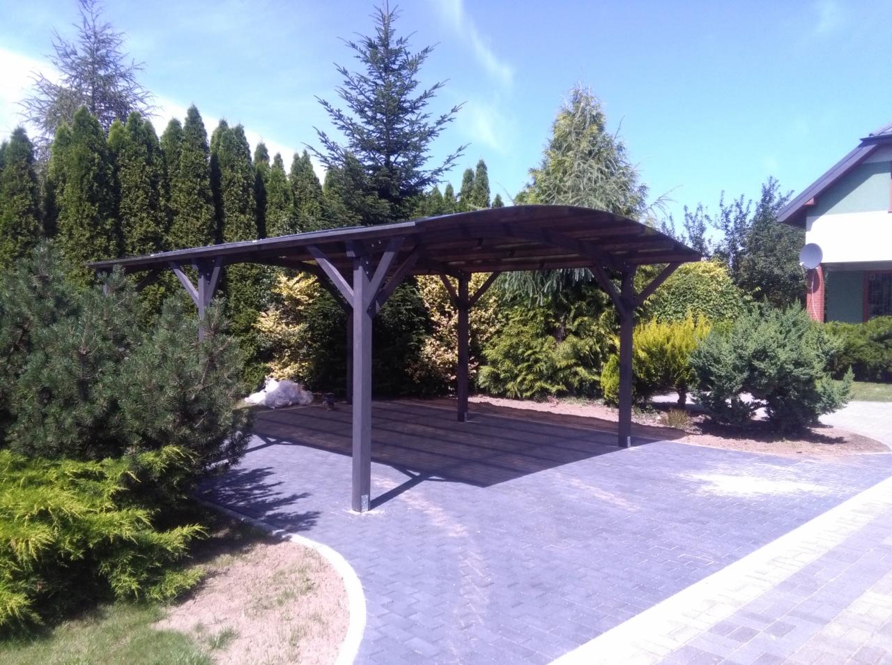 Ciemna, drewniana konstrukcja z zadaszeniem na posesji, otoczona zielenią, z widoczną kostką brukową i fragmentem budynku w tle.