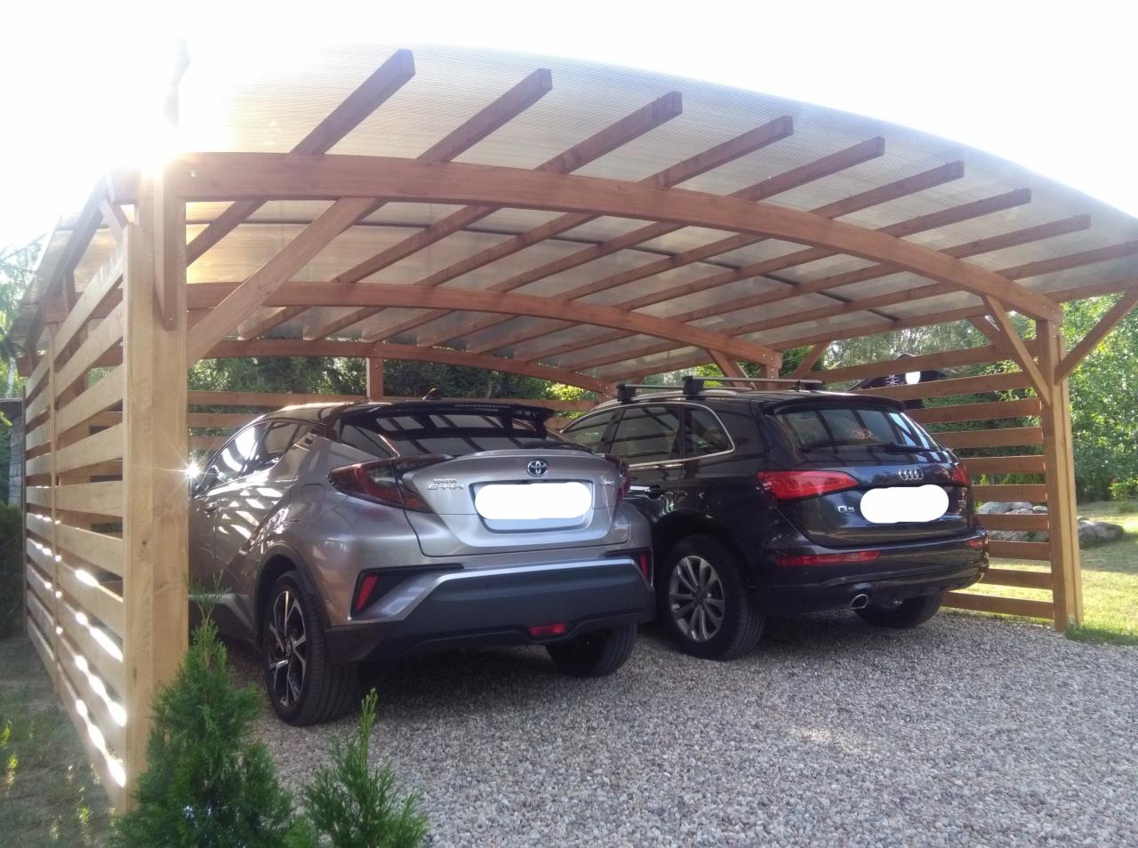 Drewniana konstrukcja z zadaszeniem z poliwęglanu, służąca jako wiata dla dwóch samochodów, stojąca na żwirowym podłożu, z widocznymi elementami ogrodzenia w tle.