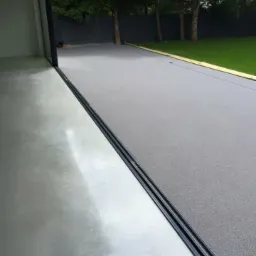 Szara, gładka powierzchnia tarasu przechodząca w betonową posadzkę wewnętrzną, oddzielona czarnym profilem dylatacyjnym, widok na ogród z drzewami i ciemnym płotem.