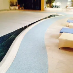 Wnętrze luksusowego basenu z częściowo odsłoniętą taflą wody, jasnoniebieska, teksturowana ścieżka wokół basenu, rattanowe meble wypoczynkowe i dekoracyjne rośliny w tle.