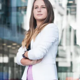 Portret kobiety w białej marynarce i różowej spódnicy, z założonymi rękami na tle nowoczesnego biurowca z szklaną fasadą.