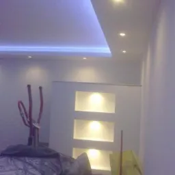 Nowoczesny sufit podwieszany z oświetleniem LED w kolorze niebieskim i punktowym, widoczne wnęki ścienne z podświetleniem, zakryty sprzęt do ćwiczeń w tle.