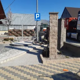 Kompozycja z różnorodnej kostki brukowej w odcieniach szarości, beżu i bieli, z elementami małej architektury ogrodowej i znakiem parkingu dla klientów w tle, widoczny wózek budowlany.