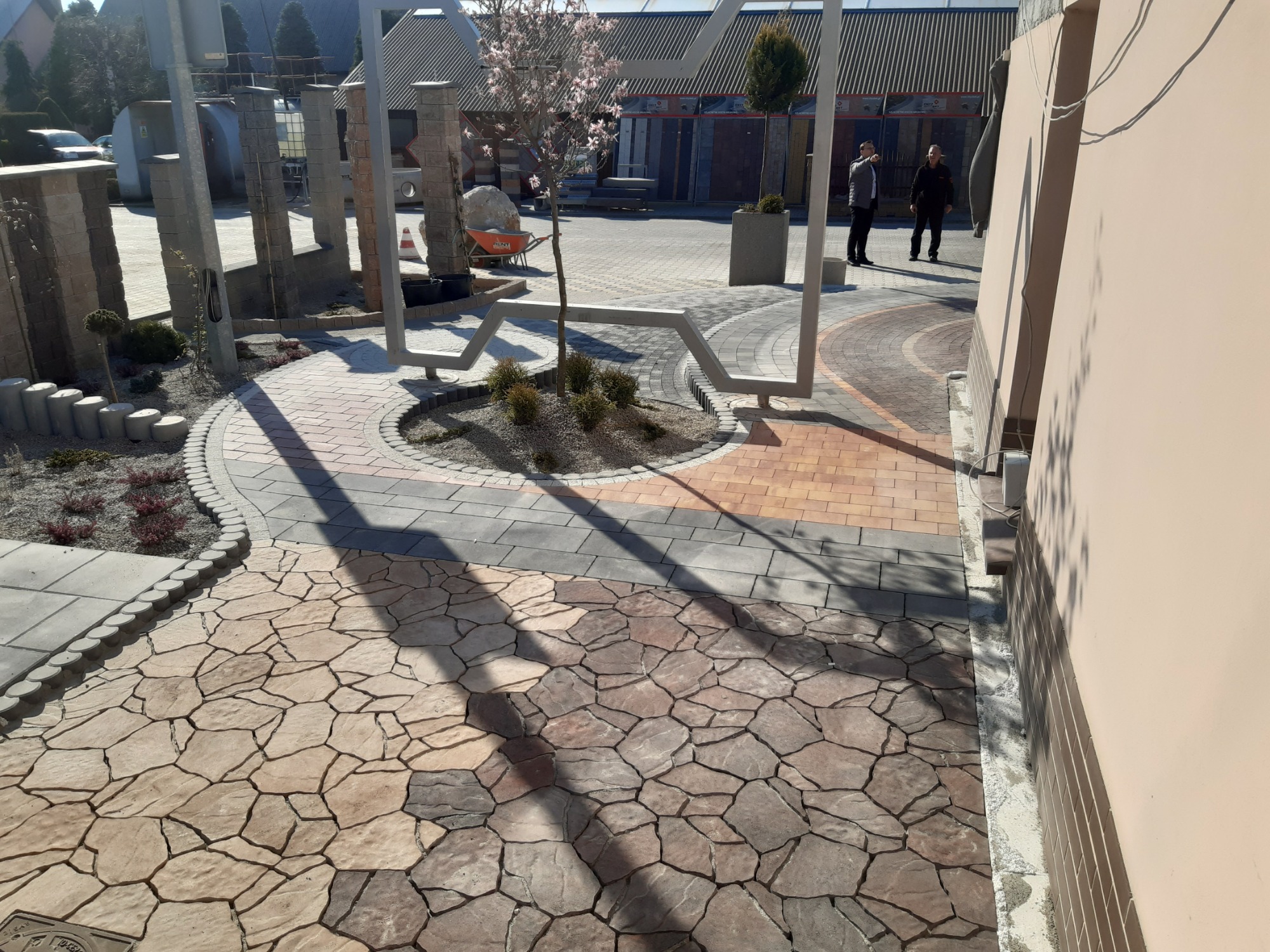 Ekspozycja różnorodnych wzorów kostki brukowej i granitowej na zewnątrz, z elementami małej architektury, takimi jak rabaty z roślinami i stalowe konstrukcje dekoracyjne, prezentowana na tle...