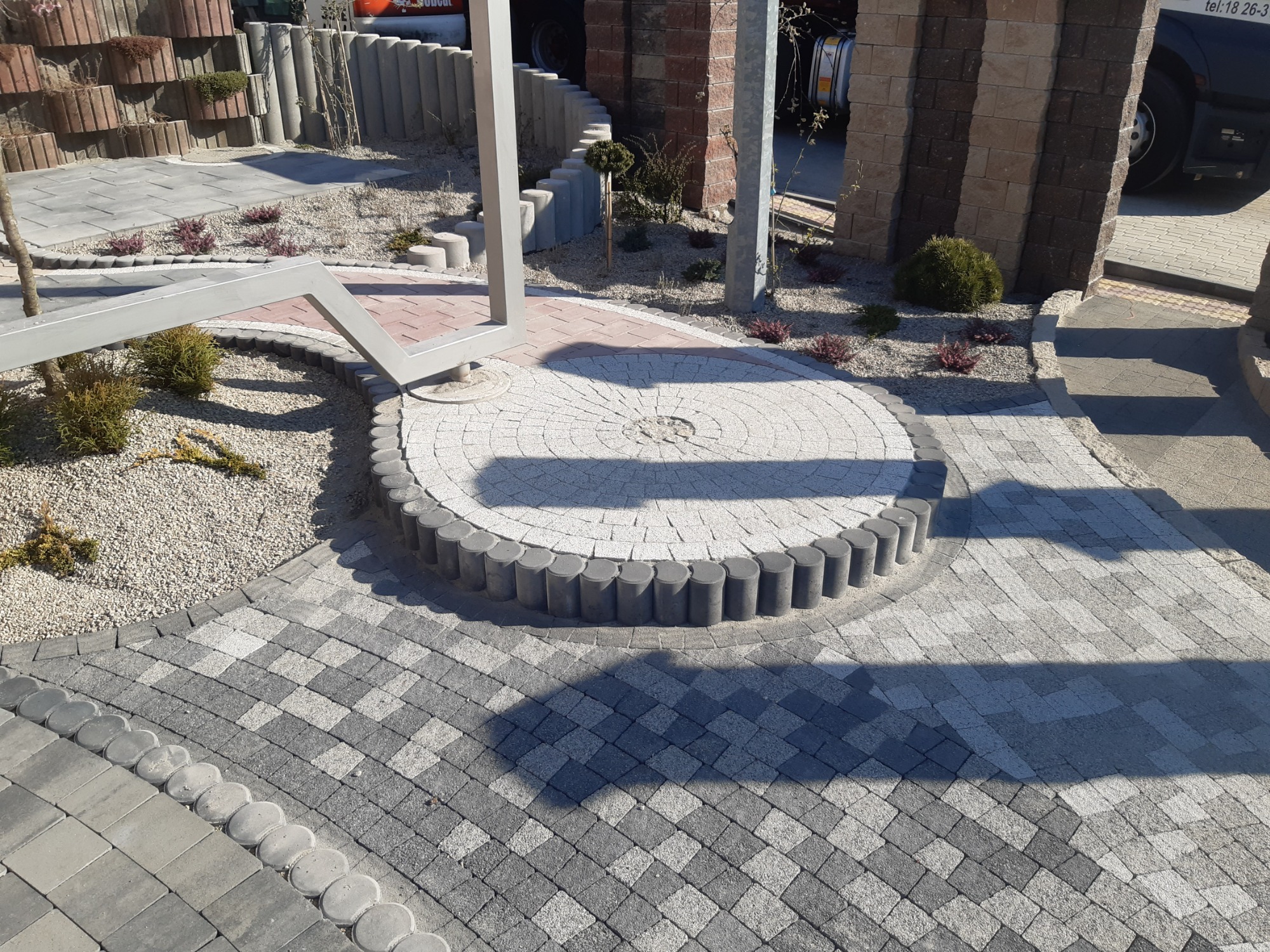 Ogród z elementami małej architektury: ścieżka z kostki brukowej w szarych odcieniach ułożona we wzór jodełki, okrągły plac z jasnej kostki otoczony obrzeżem z betonowych słupków, rabaty z krzewami...