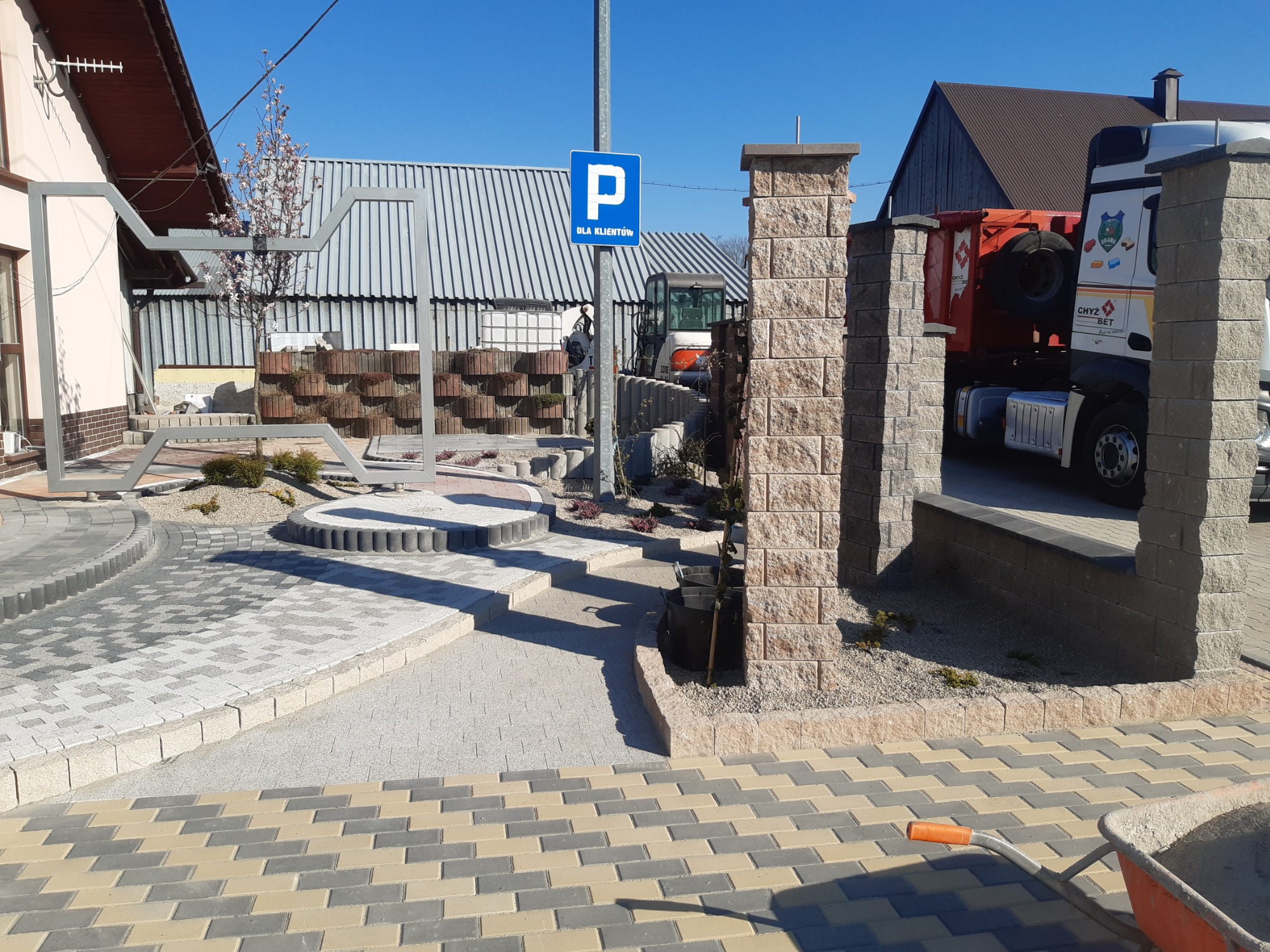 Kompozycja z różnorodnej kostki brukowej w odcieniach szarości, beżu i bieli, z elementami małej architektury ogrodowej i znakiem parkingu dla klientów w tle, widoczny wózek budowlany.