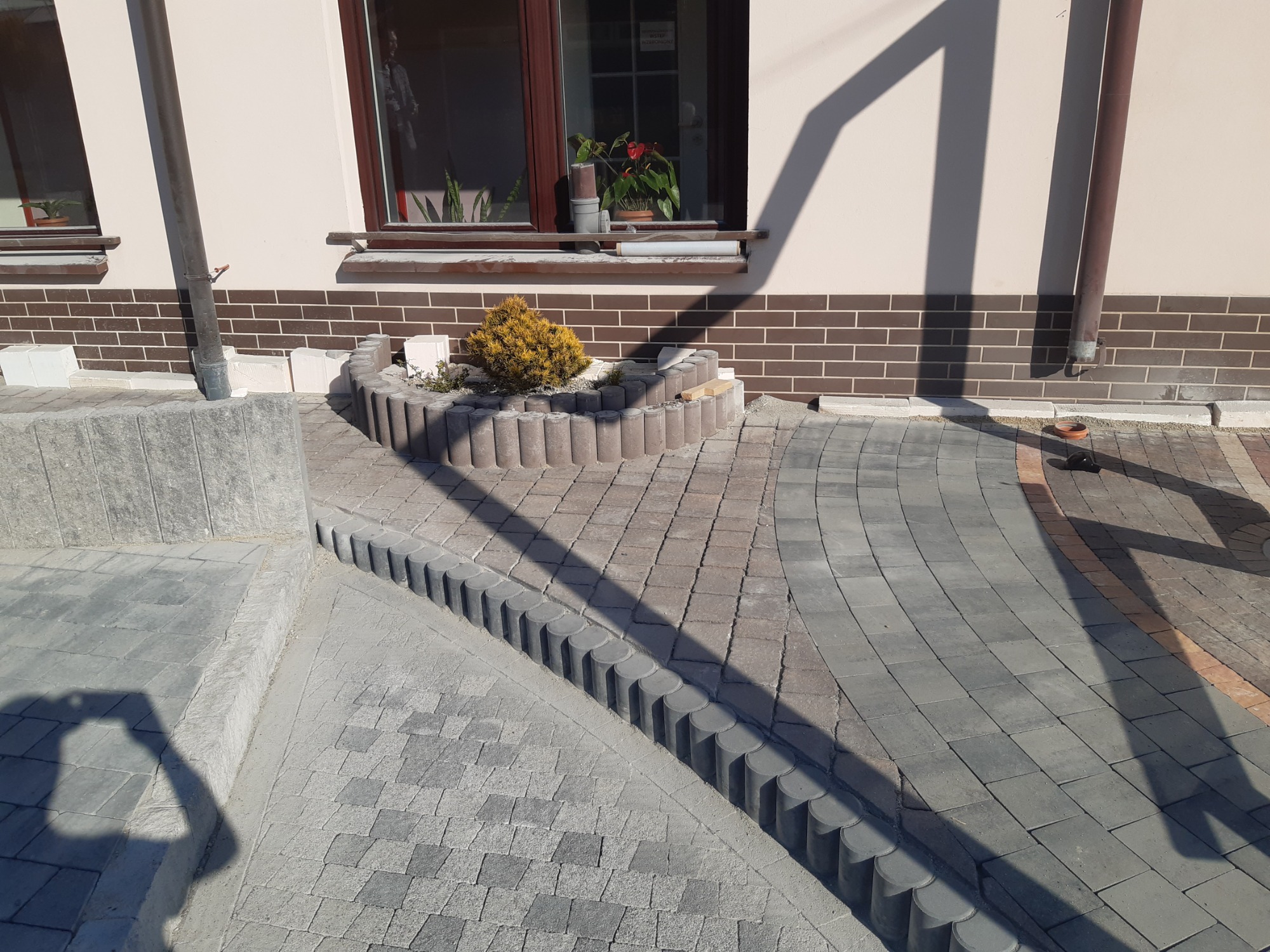 Ułożona kostka brukowa w dwóch odcieniach szarości, tworząca ścieżkę i obrzeże rabaty z rośliną iglastą, widoczny cień osoby fotografującej.