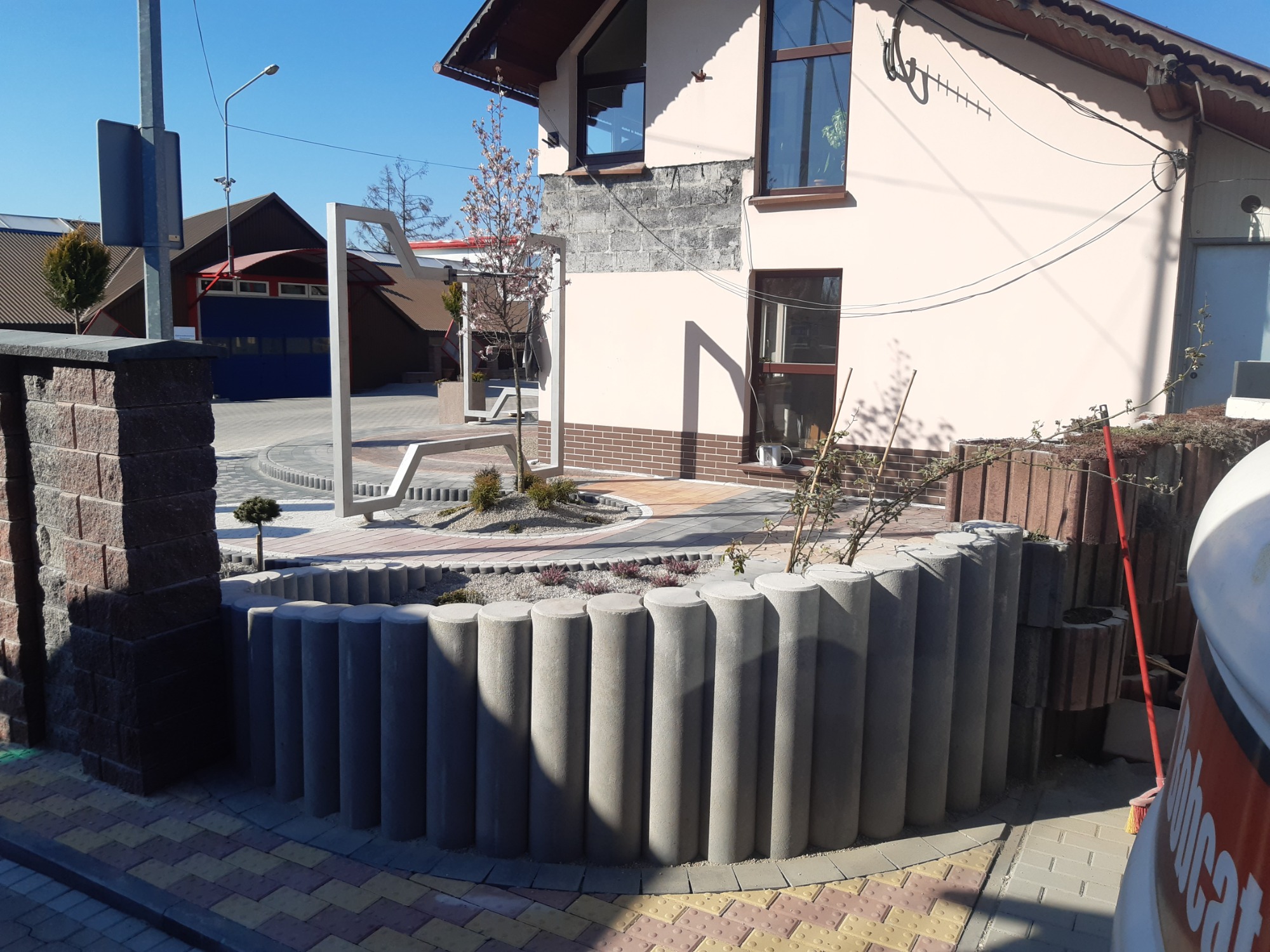 Ogródek z okrągłym obrzeżem z betonowych słupków, wyłożony jasnym żwirem i obsadzony niską roślinnością, na tle domu z kostką brukową i elementami dekoracyjnymi.