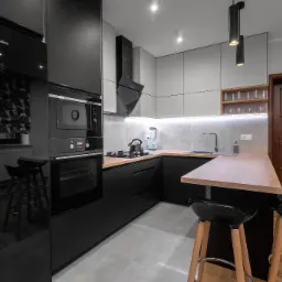 Nowoczesna kuchnia z czarnymi szafkami, drewnianymi blatami i wyspą z hokerami, oświetlona wiszącymi lampami tubowymi, z widocznym czajnikiem na płycie i blenderem w tle.