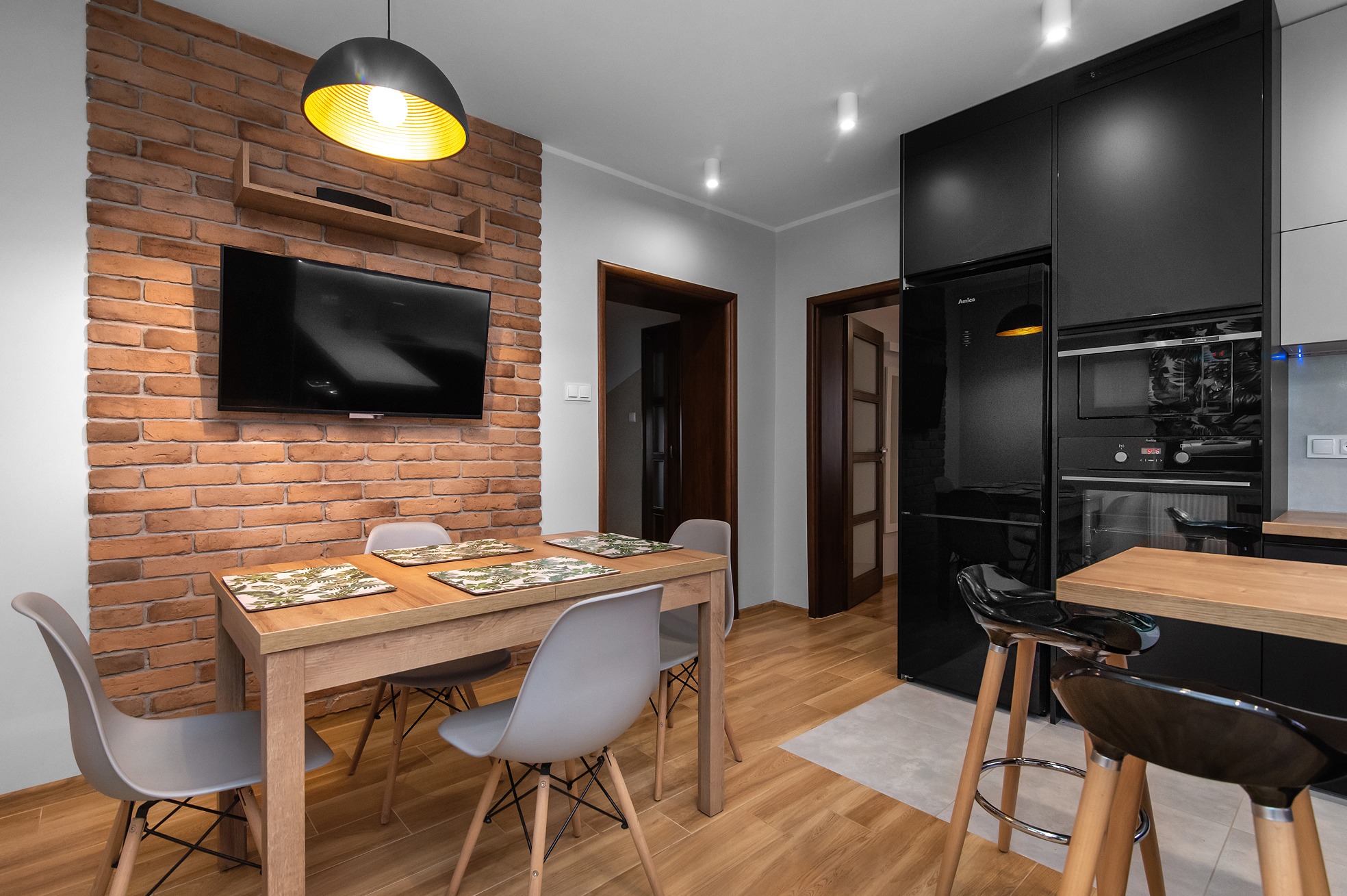 Nowoczesna jadalnia z ceglaną ścianą, drewnianym stołem i szarymi krzesłami. Nad stołem wisi czarna lampa z żółtym wnętrzem. W tle czarne szafki kuchenne i sprzęt AGD marki Amica.