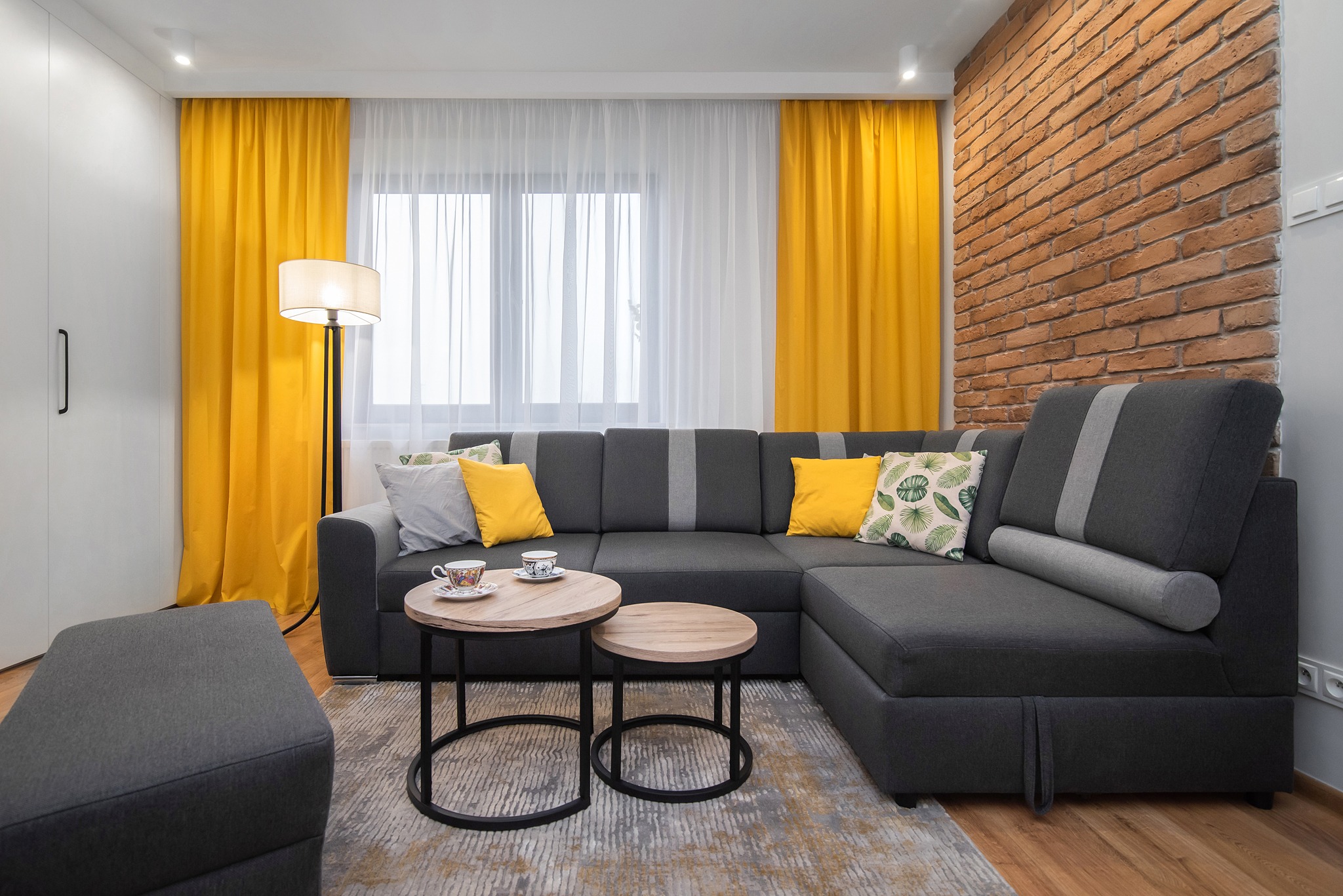 Nowoczesny salon z szarą narożną sofą, dwoma okrągłymi stolikami kawowymi, żółtymi zasłonami i ścianą z cegły, dopełniony lampą podłogową i poduszkami dekoracyjnymi.