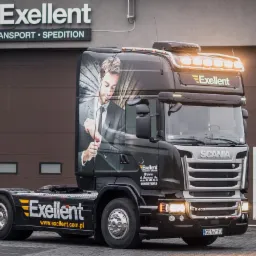 Czarny ciągnik siodłowy Scania z grafiką mężczyzny w garniturze na kabinie, logo firmy Exellent Transport Spedition na boku i dachu, zaparkowany przed budynkiem firmy z brązową bramą garażową.