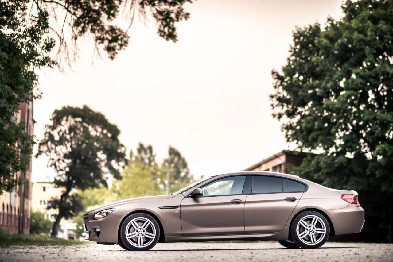 Bursztynowy sedan BMW serii 6 z aluminiowymi felgami, sfotografowany z boku na tle zieleni i budynków.