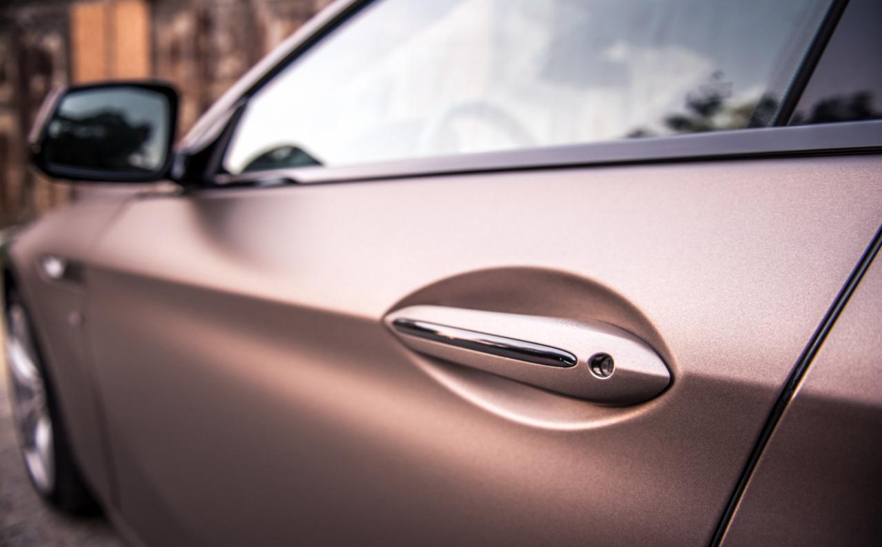 Matowy, brązowo-złoty lakier na drzwiach samochodu, z chromowaną klamką i odbiciem otoczenia w szybie.