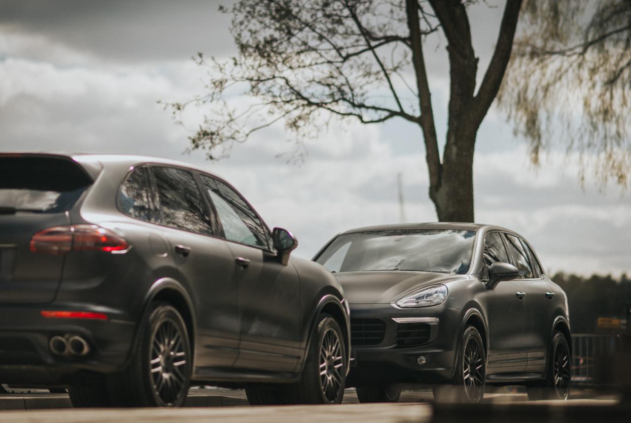 Dwa matowe, grafitowe samochody Porsche Cayenne zaparkowane obok siebie na tle pochmurnego nieba i drzewa bez liści.