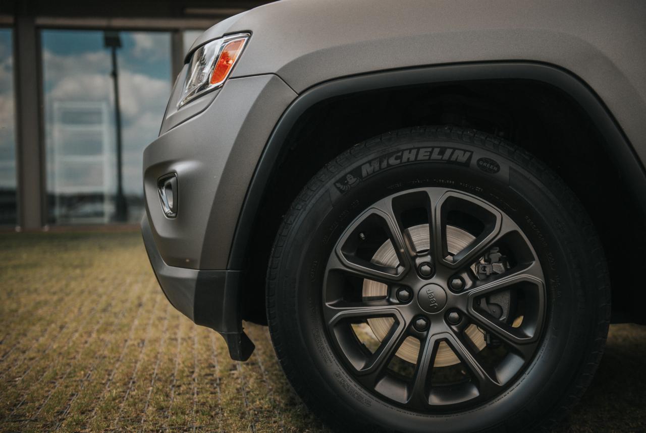 Szary Jeep Grand Cherokee z czarnymi felgami i oponami Michelin, zaparkowany na trawniku przed nowoczesnym budynkiem z dużymi oknami.